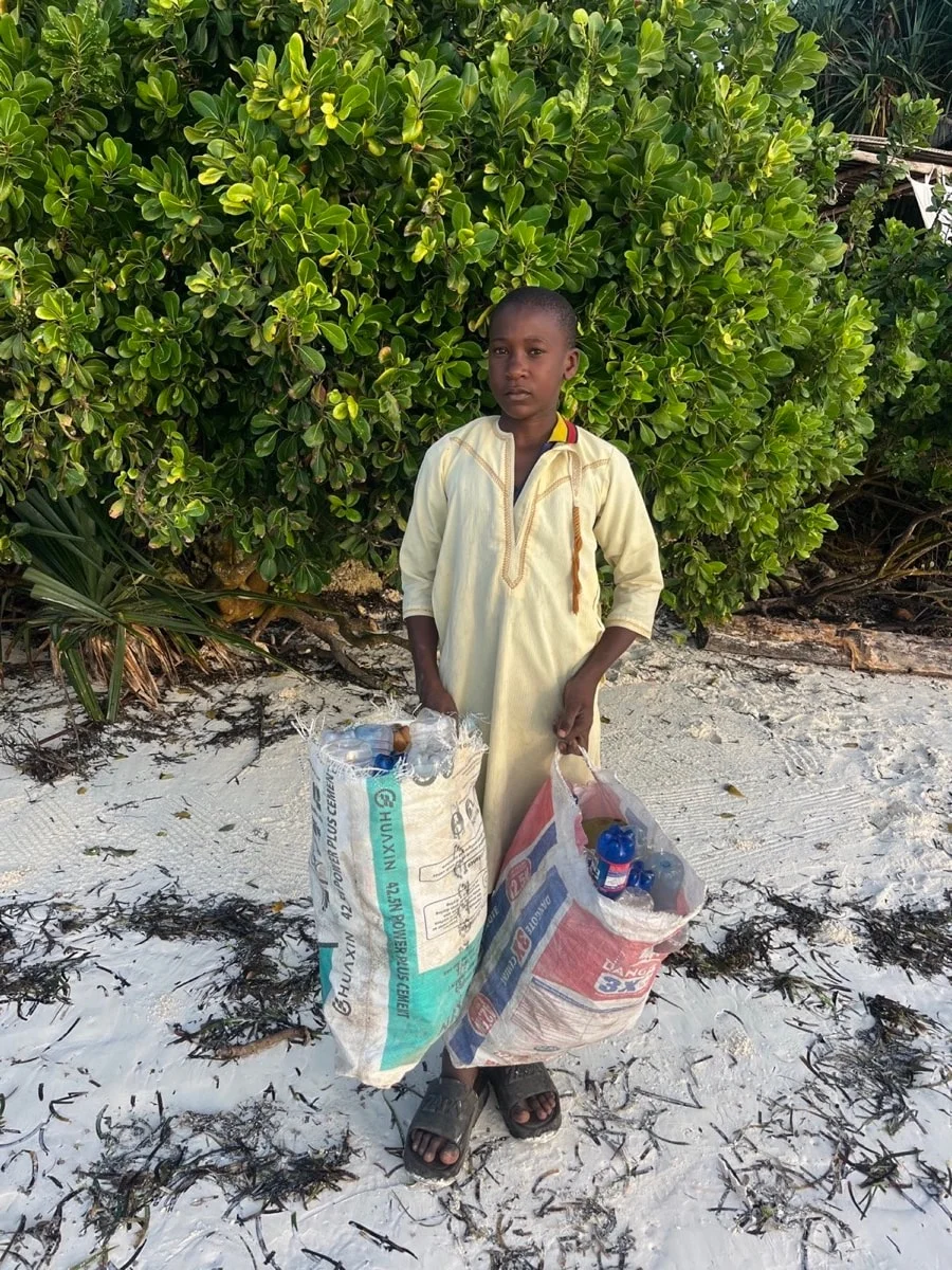 A young local boy holding two sacks of collected plastic bottles on a white-sand Zanzibar beach